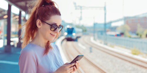 Eine Frau steht am Gleis und schaut lächelnd auf ihr Handy.