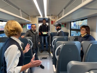 Menschen stehen in einem Zug vor den Sitzplätzen und agieren miteinander.